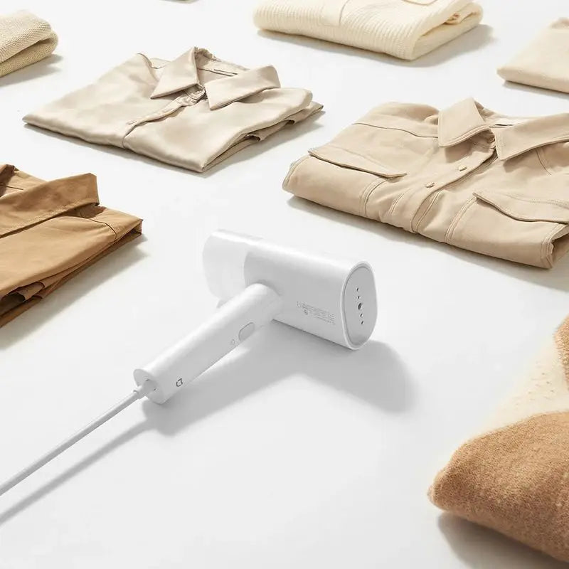 White handheld steamer on a light surface with folded beige clothing items.